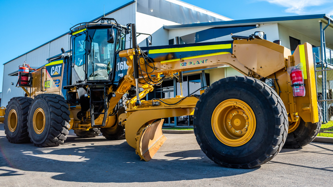 Yellow earthmoving equipment, grader, stationery outside a building.
